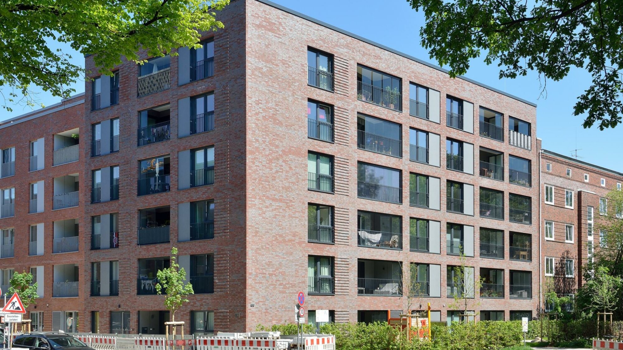 Großes, modernes Wohngebäude mit roter Klinkerfassade und vielen Balkonen an einer Straßenecke; im Vordergrund Autos, Baustellenabsperrungen und frisch gepflanzte Bäume, blauer Himmel im Hintergrund.