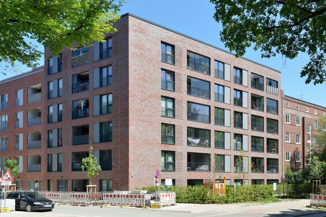 Großes, modernes Wohngebäude mit roter Klinkerfassade und vielen Balkonen an einer Straßenecke; im Vordergrund Autos, Baustellenabsperrungen und frisch gepflanzte Bäume, blauer Himmel im Hintergrund.