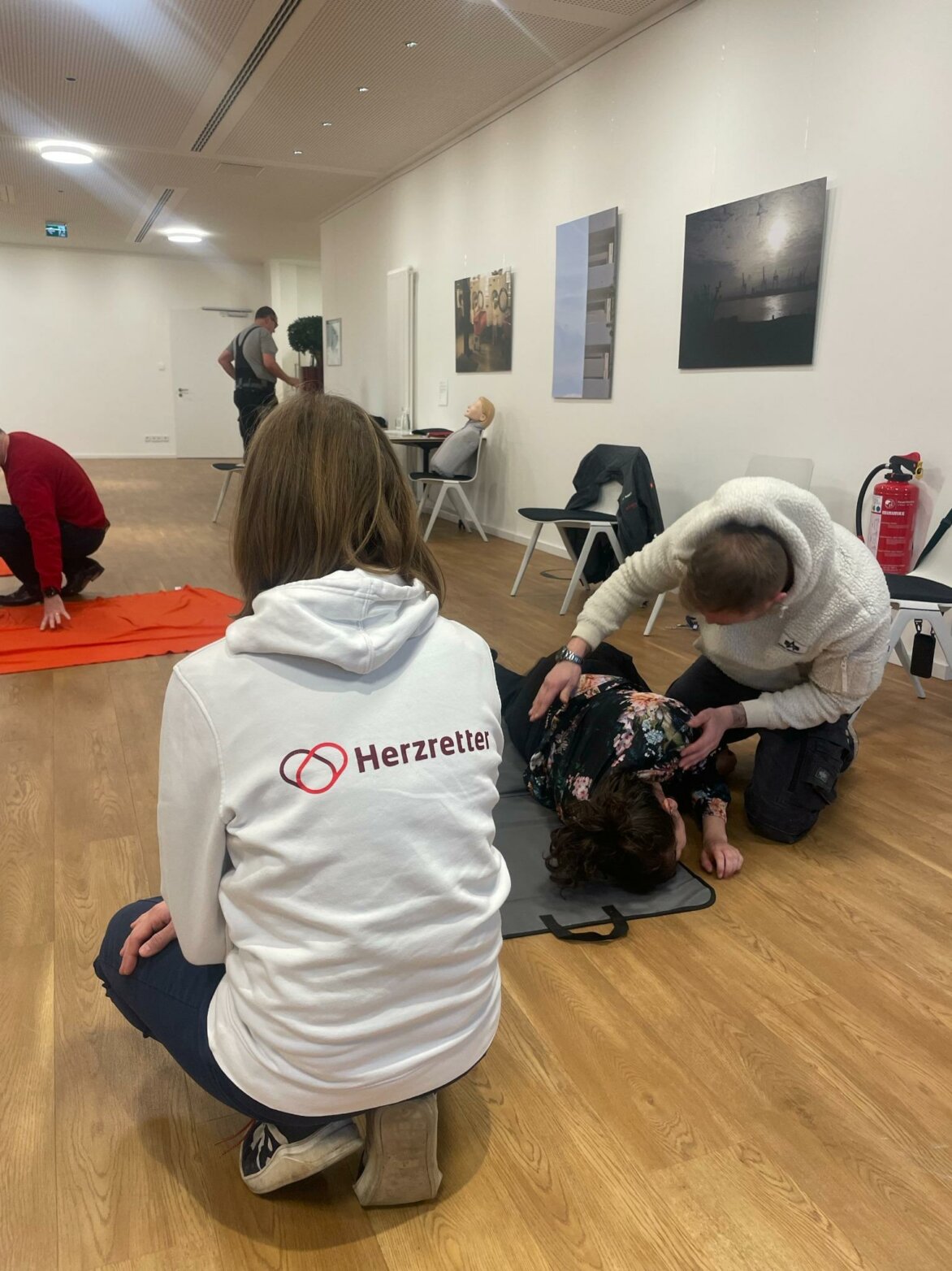 Eine Person in weißem Hoodie mit der Aufschrift „Herzretter“ beobachtet in einem Schulungsraum eine Übung zur stabilen Seitenlage, bei der eine andere Person eine auf dem Boden liegende Teilnehmerin lagestabil betreut; im Hintergrund sind weitere Übungssituationen mit Erste-Hilfe-Puppen zu sehen.