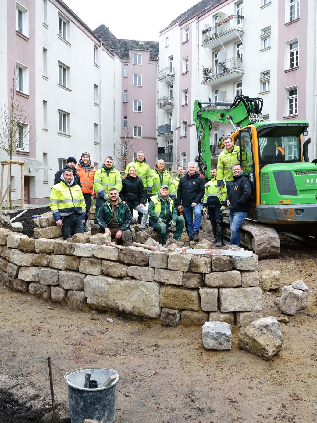 Gruppe von etwa 16 Personen, größtenteils in Arbeitskleidung, steht und sitzt vor einer neu errichteten Trockenmauer aus Naturstein in einem Innenhof zwischen mehrstöckigen Wohngebäuden; im Hintergrund ein grüner Bagger.
