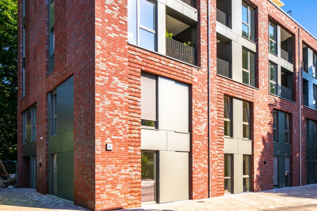 Neubau eines mehrstöckigen Wohngebäudes mit roter Klinkerfassade und großen, modernen Fenster- und Balkonelementen; gepflasterter Gehweg und Straße im Vordergrund, Bäume im Hintergrund.