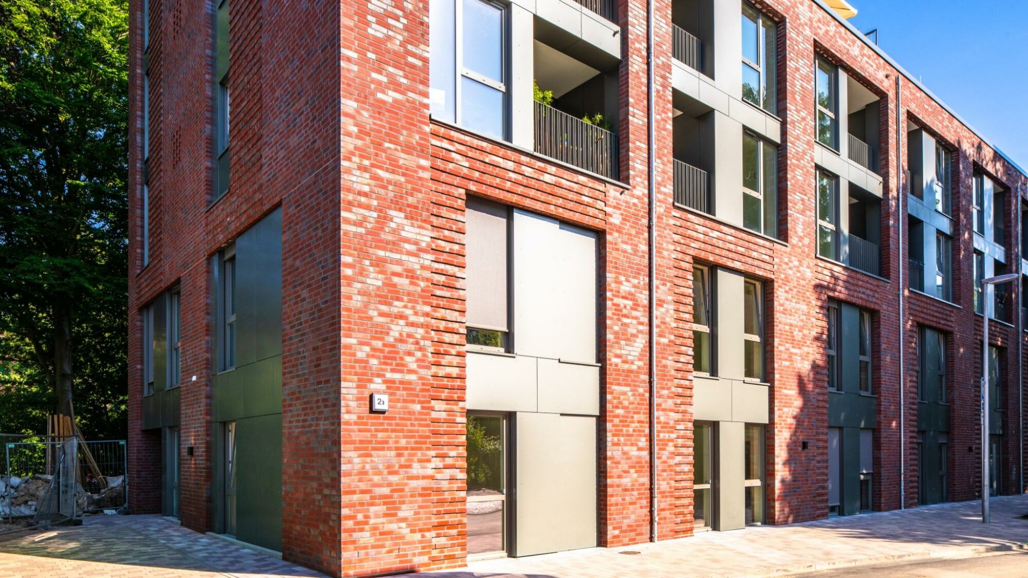 Neubau eines mehrstöckigen Wohngebäudes mit roter Klinkerfassade und großen, modernen Fenster- und Balkonelementen; gepflasterter Gehweg und Straße im Vordergrund, Bäume im Hintergrund.