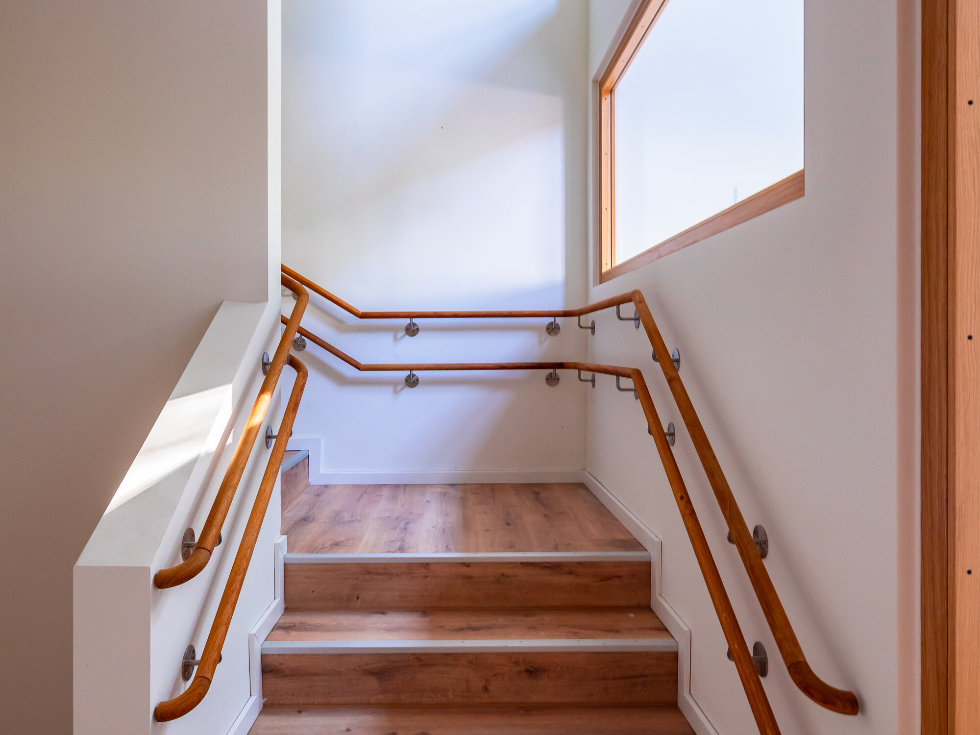Innenaufnahme eines Treppenhauses mit sechs Holzstufen, beidseitigen Handläufen in unterschiedlichen Höhen und einem großen Fenster im oberen Bereich; die Wände und Decke sind weiß, der Boden besteht aus Holzlaminat.