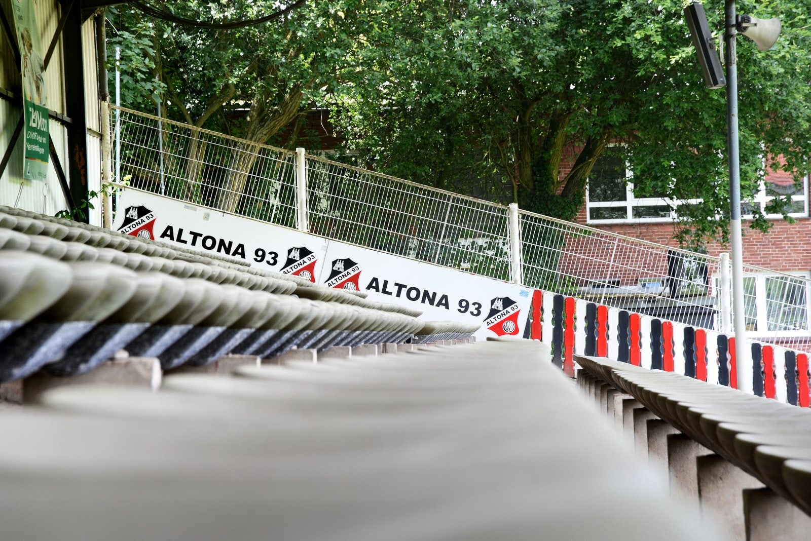 Leere Tribünenplätze im Adolf-Jäger-Stadion mit Blick auf Werbebande von Altona 93; im Hintergrund Zaun, Bäume und ein Gebäude mit weißen Fenstern; Vereinsfarben Rot, Schwarz und Weiß sind mehrfach vertreten.