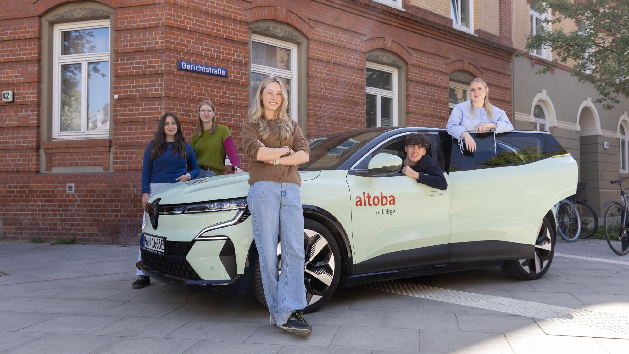 Fünf junge Erwachsene stehen und sitzen um ein hellgrünes Auto mit altoba-Logo vor einem Backsteingebäude an der Ecke Haubachstraße/Gerichtstraße; es wirkt wie eine Azubi-Präsentation.
