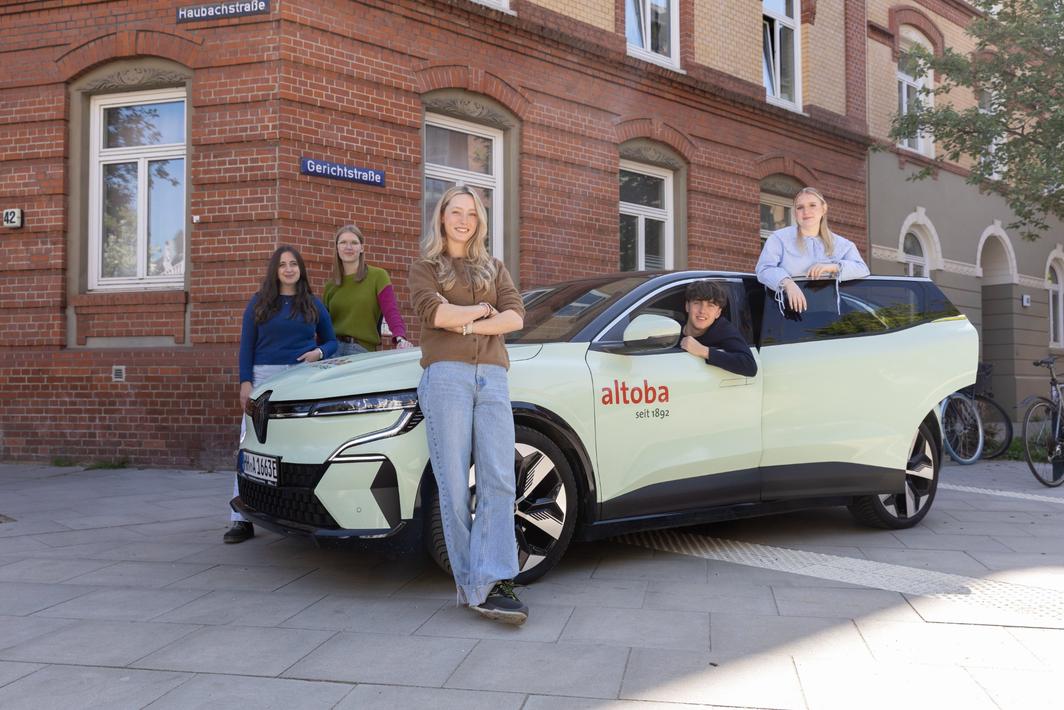 Fünf junge Erwachsene stehen und sitzen um ein hellgrünes Auto mit altoba-Logo vor einem Backsteingebäude an der Ecke Haubachstraße/Gerichtstraße; es wirkt wie eine Azubi-Präsentation.