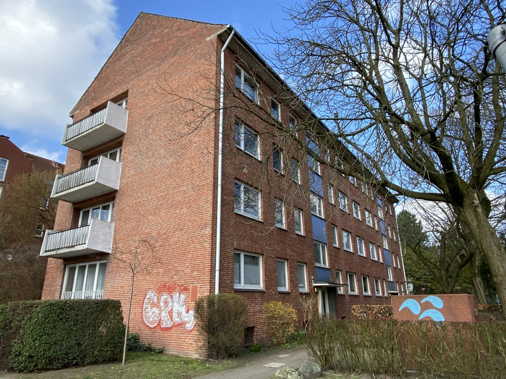 Viergeschossiges Wohnhaus aus rotem Klinker mit Balkonen an der Stirnseite und großflächigem Efeubewuchs; Gehweg und dichte Bepflanzung im Vordergrund, teilweise belaubter Baum rechts im Bild.