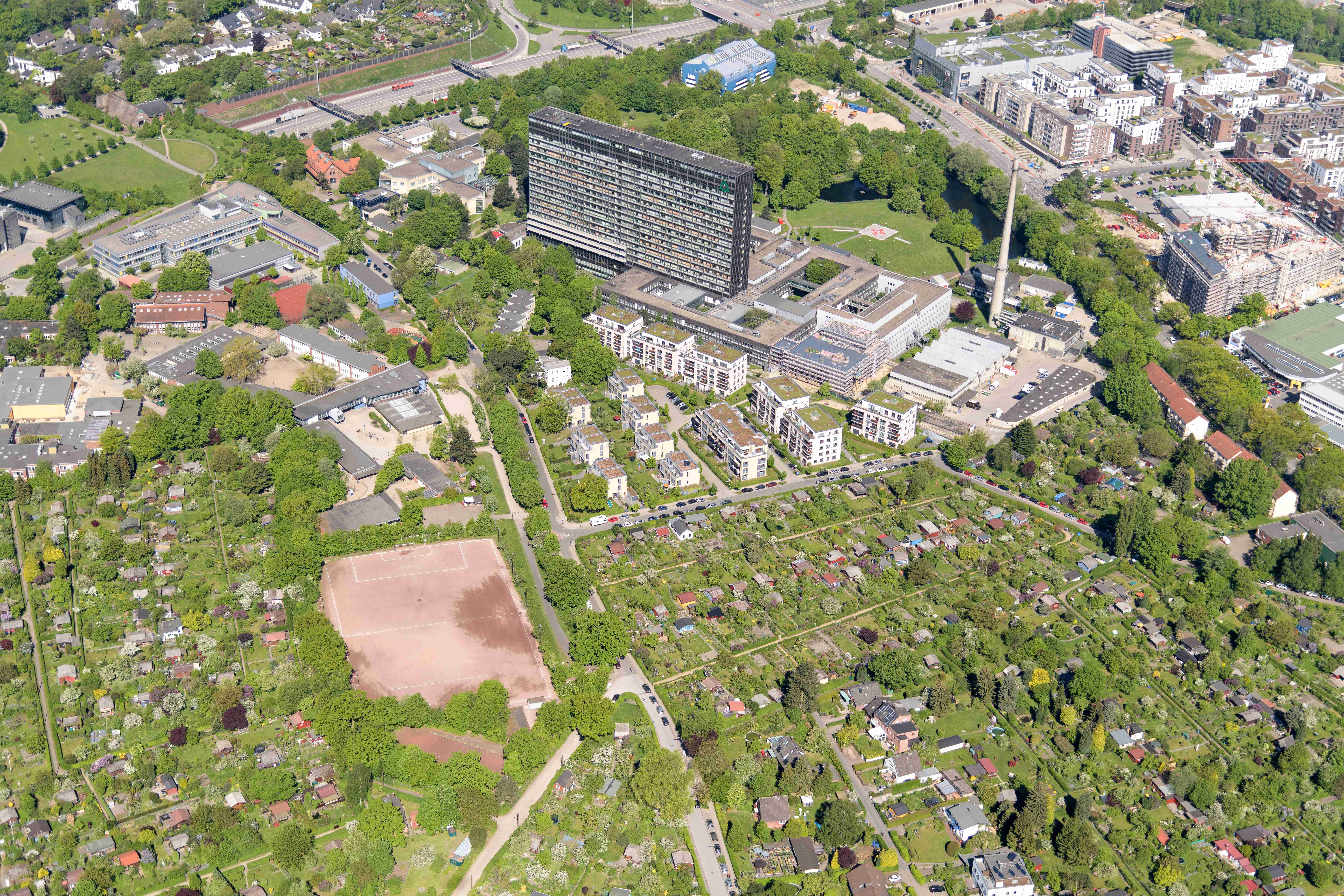 Luftaufnahme eines Stadtteils mit Wohnhäusern, Kleingartenanlagen, Sportplätzen und vielen Bäumen; im Vordergrund moderne Wohngebäude, im Hintergrund dicht bebaute Stadtstruktur.