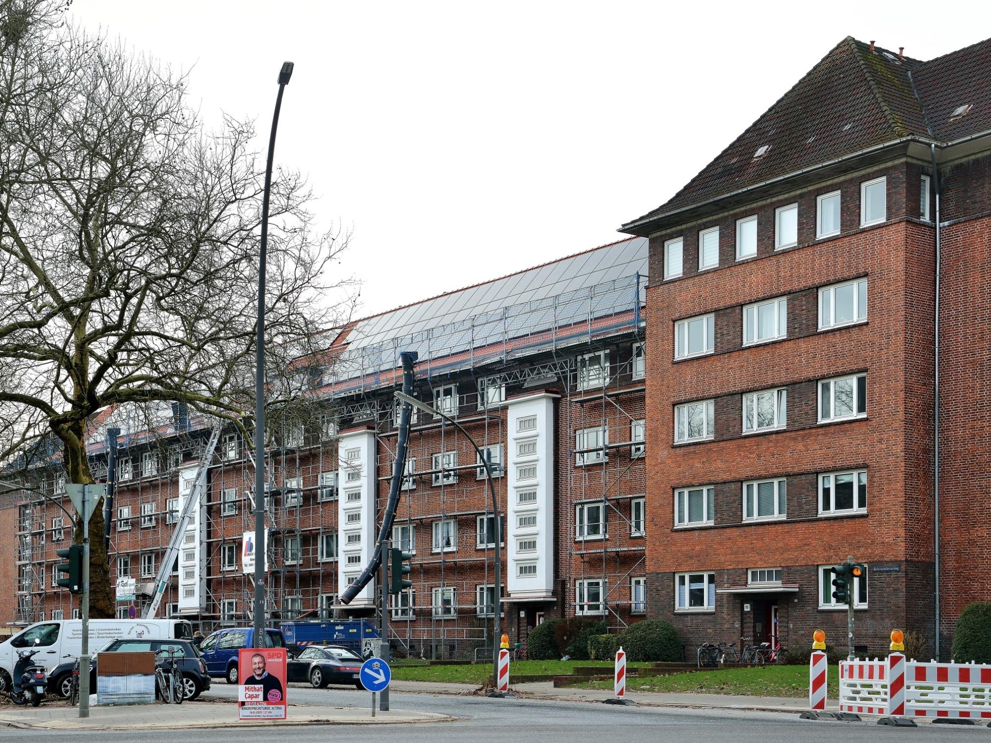 Mehrstöckiges Backsteingebäude an einer Straßenecke mit eingerüstetem Dachbereich, auf dem Arbeiten für eine Photovoltaikanlage stattfinden; davor Straßenverkehr, Bauabsperrungen und ein Wahlplakat.