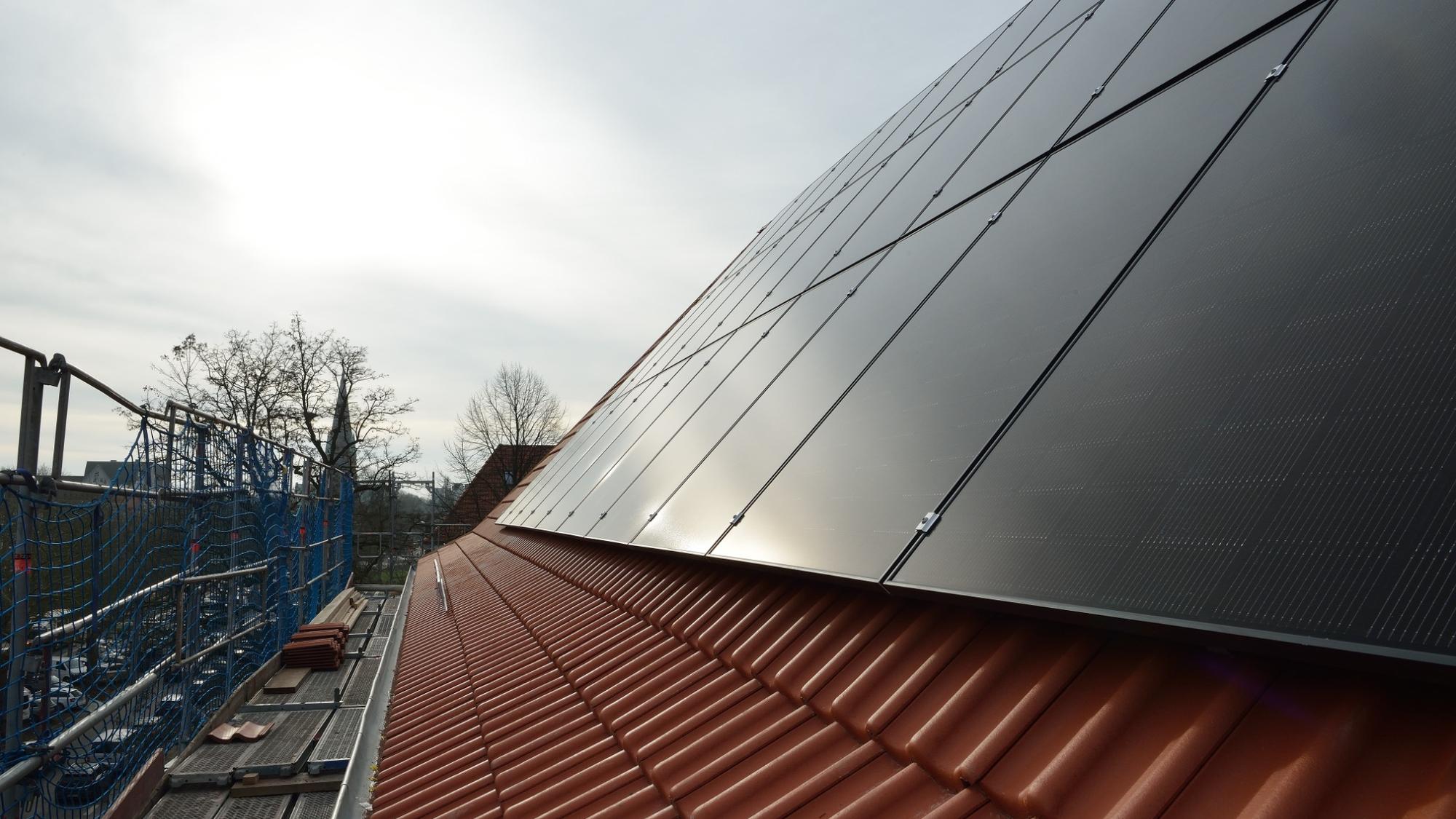 Nahaufnahme einer Dachfläche mit dunkel glänzenden Photovoltaikmodulen auf roten Ziegeln; links ist ein Baugerüst mit Sicherungsnetz und gestapelten Dachziegeln zu sehen, im Hintergrund stehen kahle Bäume und ein Kirchturm.
