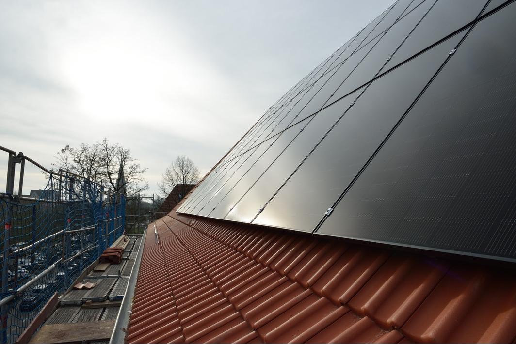 Nahaufnahme einer Dachfläche mit dunkel glänzenden Photovoltaikmodulen auf roten Ziegeln; links ist ein Baugerüst mit Sicherungsnetz und gestapelten Dachziegeln zu sehen, im Hintergrund stehen kahle Bäume und ein Kirchturm.