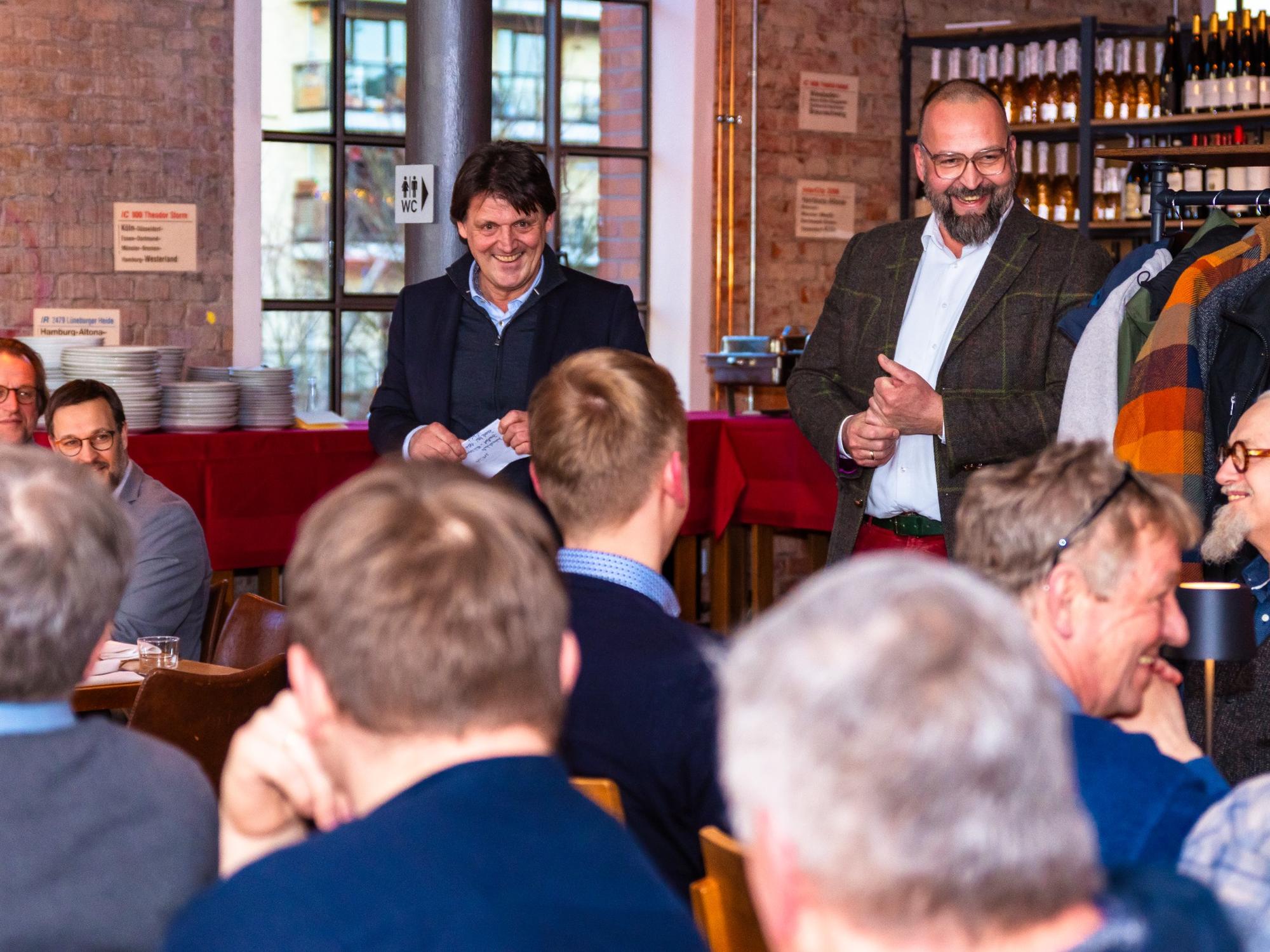 Gruppe von etwa zehn Männern in einem Restaurant oder Veranstaltungsraum; zwei Männer stehen lächelnd im Vordergrund und sprechen zu den anderen, die an Tischen sitzen und zuhören; im Hintergrund Regale mit Weinflaschen und Gläsern, rote Tischdecken und ein Stapel Teller sichtbar; freundliche, gesellige Stimmung.