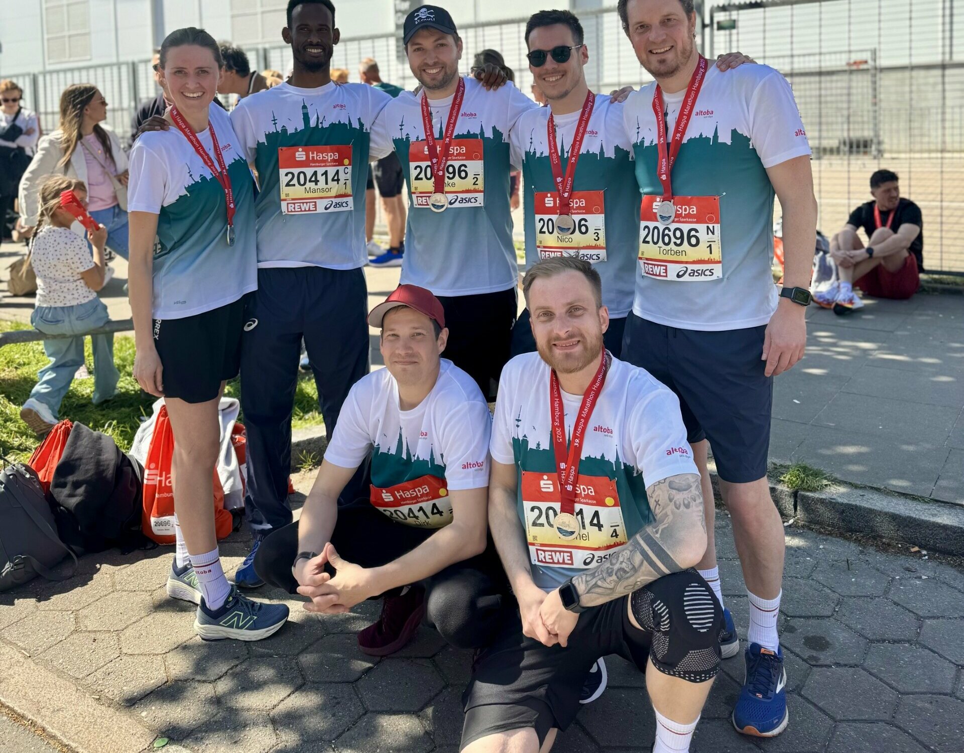 Sechs Männer und eine Frau in Laufkleidung mit altoba-Shirts und Startnummern stehen und knien lächelnd vor einer Messehalle, alle tragen Medaillen vom Haspa Marathon 2025