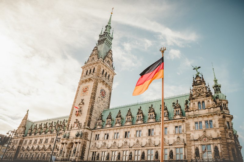 Historisches Rathaus in Hamburg mit grünem Kupferdach und Turmuhr, davor eine wehende Deutschlandfahne bei leicht bewölktem Himmel.