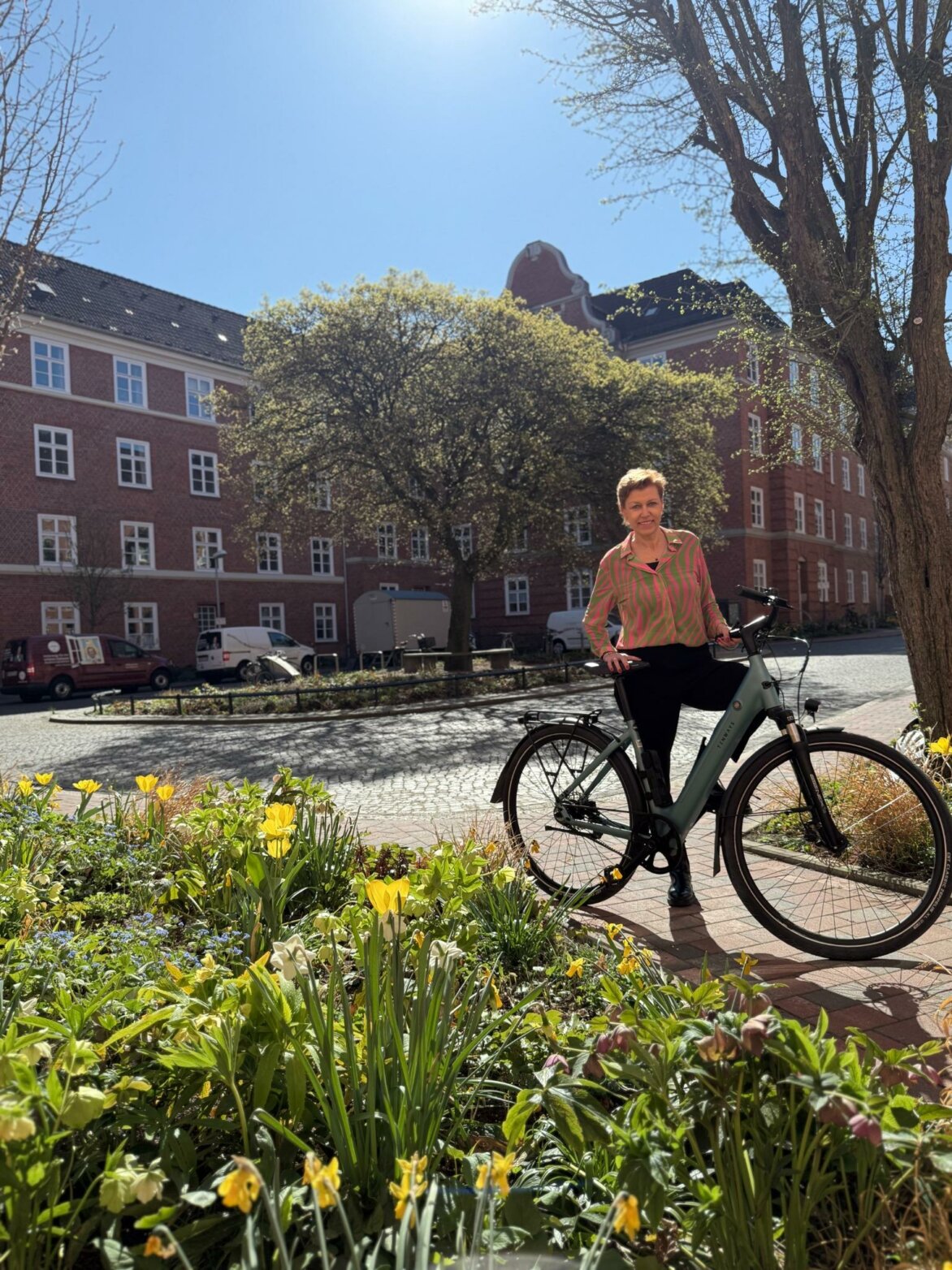 Eine Frau mit bunter Bluse steht bei Sonnenschein mit einem Fahrrad auf einem Weg vor einem blühenden Blumenbeet; im Hintergrund ein historisches Backsteingebäude mit weißen Fenstern.