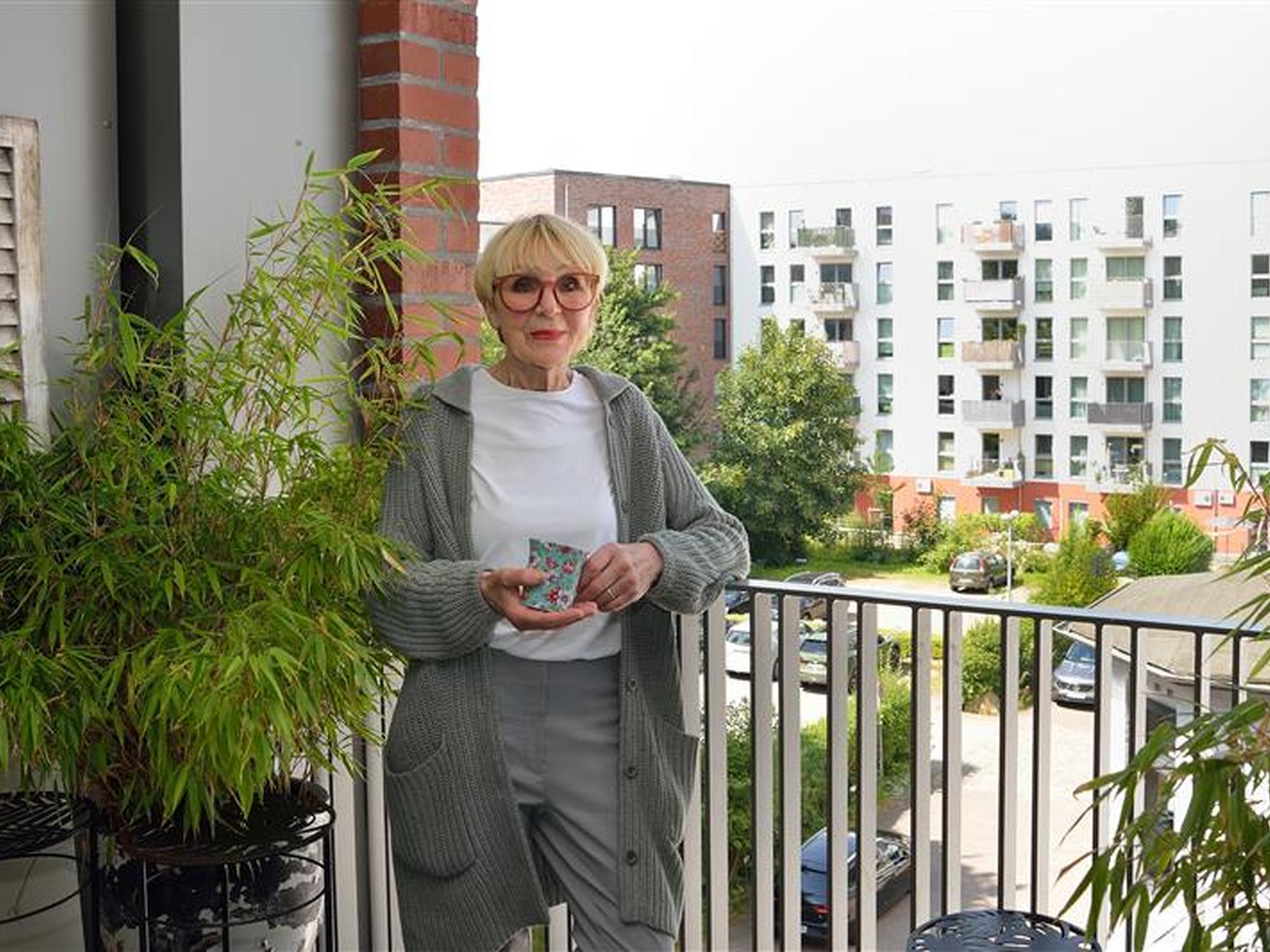 Ältere Frau mit blondem Kurzhaarschnitt und Brille steht auf einem Balkon, hält eine bunte Tasse in beiden Händen und lächelt leicht; sie trägt eine graue Strickjacke, ein weißes T-Shirt und graue Hose. Im Hintergrund sind moderne mehrstöckige Wohngebäude, Bäume und geparkte Autos zu sehen. Auf dem Balkon stehen grüne Pflanzen in schwarzen Töpfen.