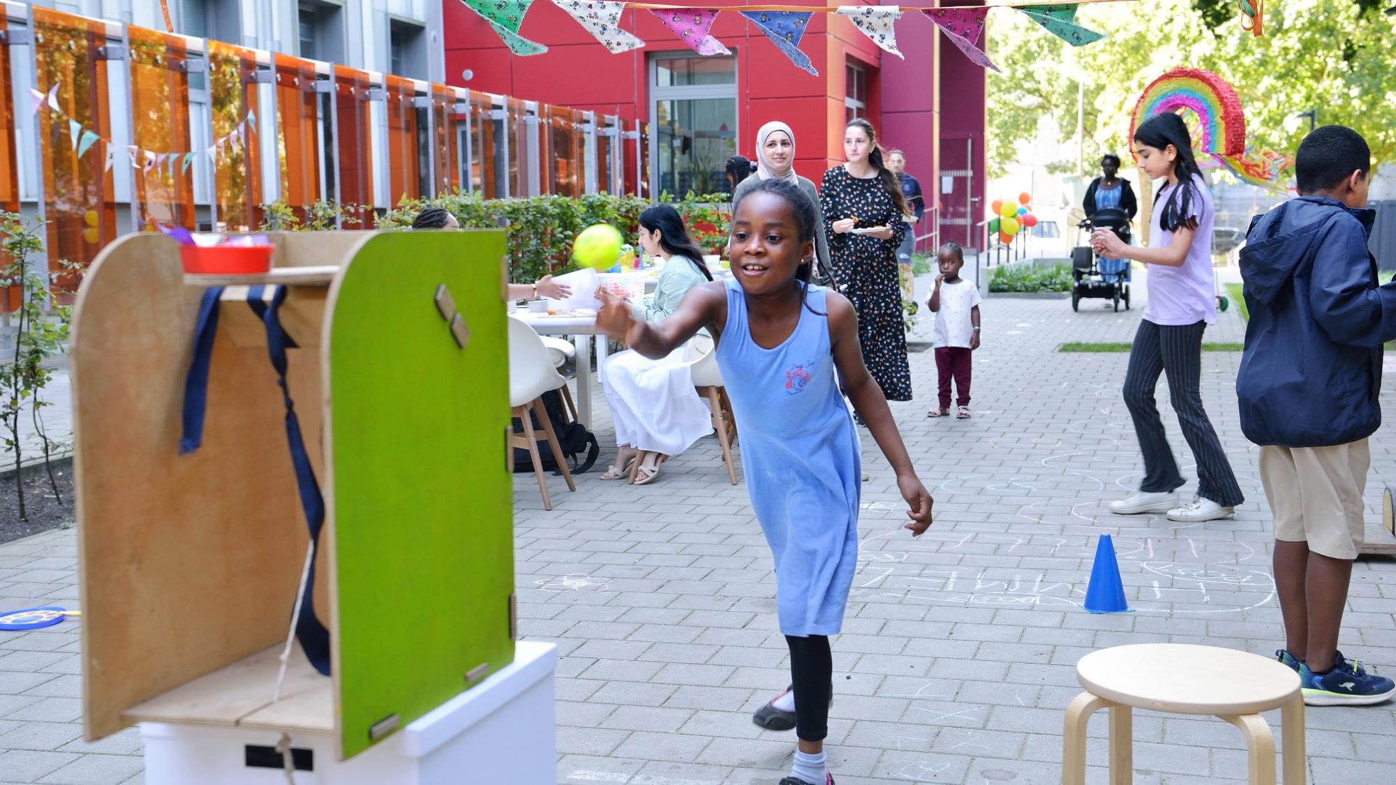 Fröhliches Straßenspiel mit Kindern und Erwachsenen bei einem Nachbarschaftsfest; im Vordergrund wirft ein Mädchen in blauer Kleidung einen Ball auf ein Wurfspiel, im Hintergrund bunte Wimpelketten, Luftballons und weitere spielende Kinder vor einem modernen Wohngebäude.