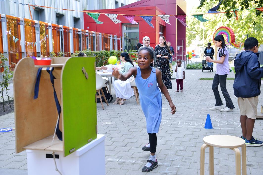 Fröhliches Straßenspiel mit Kindern und Erwachsenen bei einem Nachbarschaftsfest; im Vordergrund wirft ein Mädchen in blauer Kleidung einen Ball auf ein Wurfspiel, im Hintergrund bunte Wimpelketten, Luftballons und weitere spielende Kinder vor einem modernen Wohngebäude.