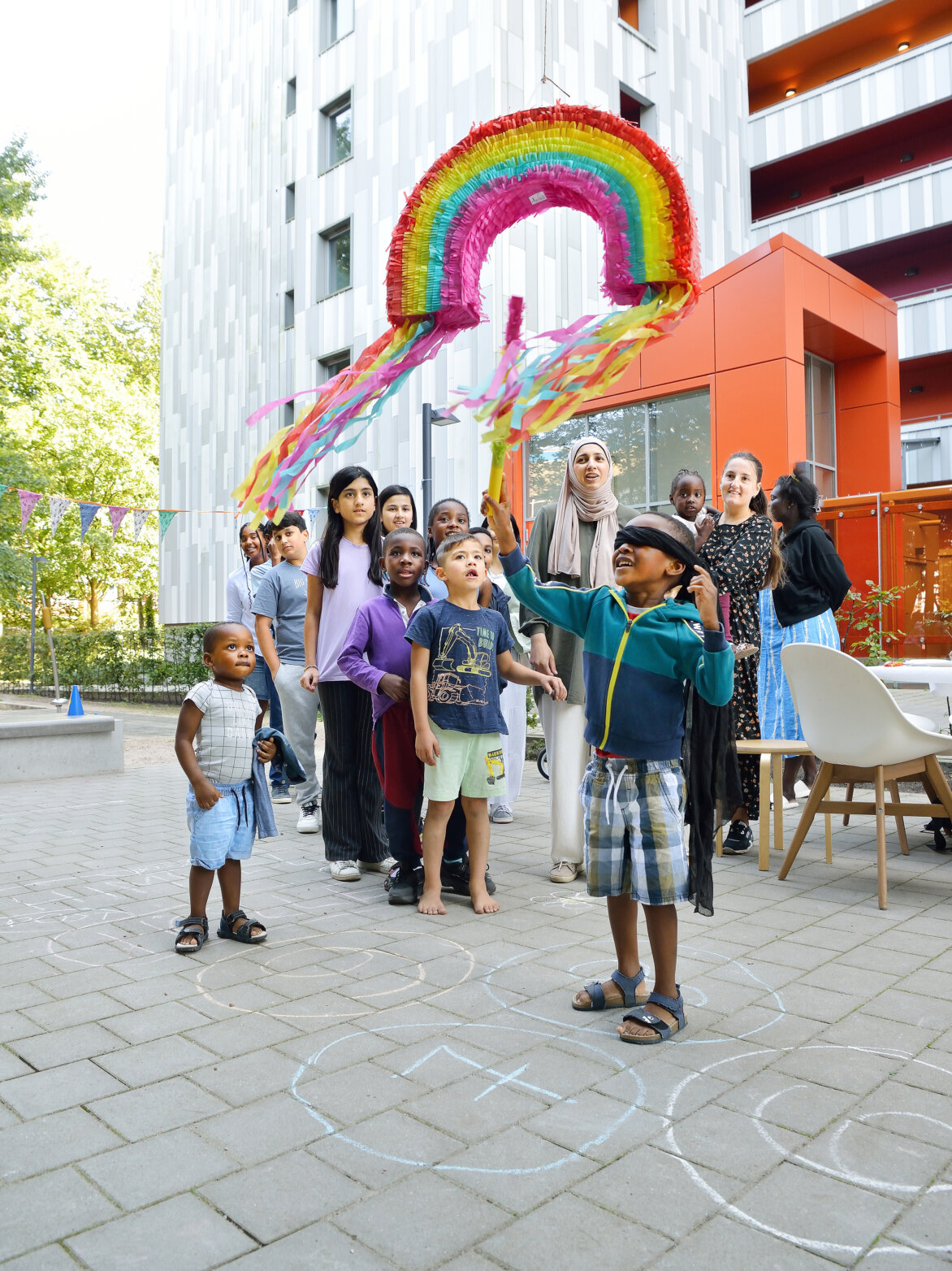 Ein Junge mit verbundenen Augen schlägt mit einem Stock auf eine bunte Regenbogen-Piñata, während andere Kinder und einige Erwachsene auf einem Hof vor einem modernen Wohngebäude zuschauen.