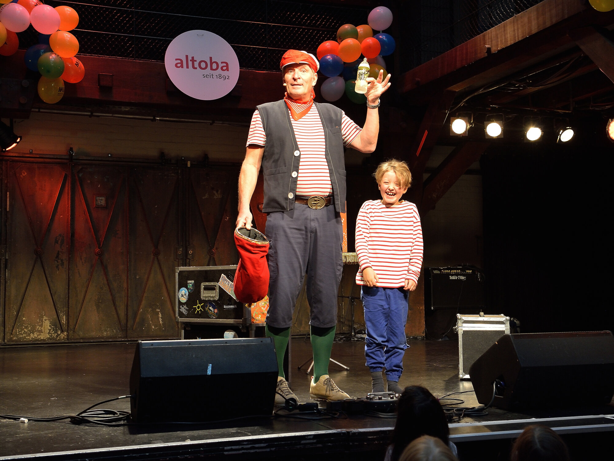 Ein Künstler in maritimer Kleidung steht mit einem lachenden Kind auf einer Bühne bei einer altoba-Veranstaltung; beide tragen gestreifte Shirts, im Hintergrund bunte Luftballons und ein altoba-Logo.