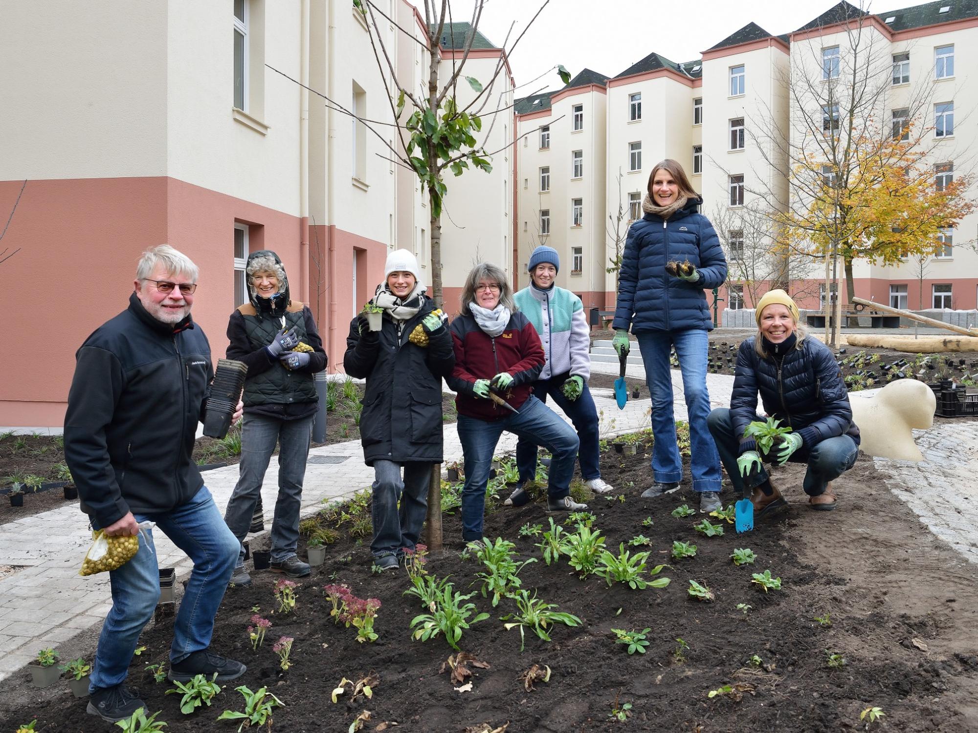 Acht Personen unterschiedlichen Alters pflanzen gemeinsam Jungpflanzen in ein Beet im Innenhof einer sanierten Wohnanlage; im Hintergrund helle Mehrfamilienhäuser und herbstlich gefärbte Bäume.