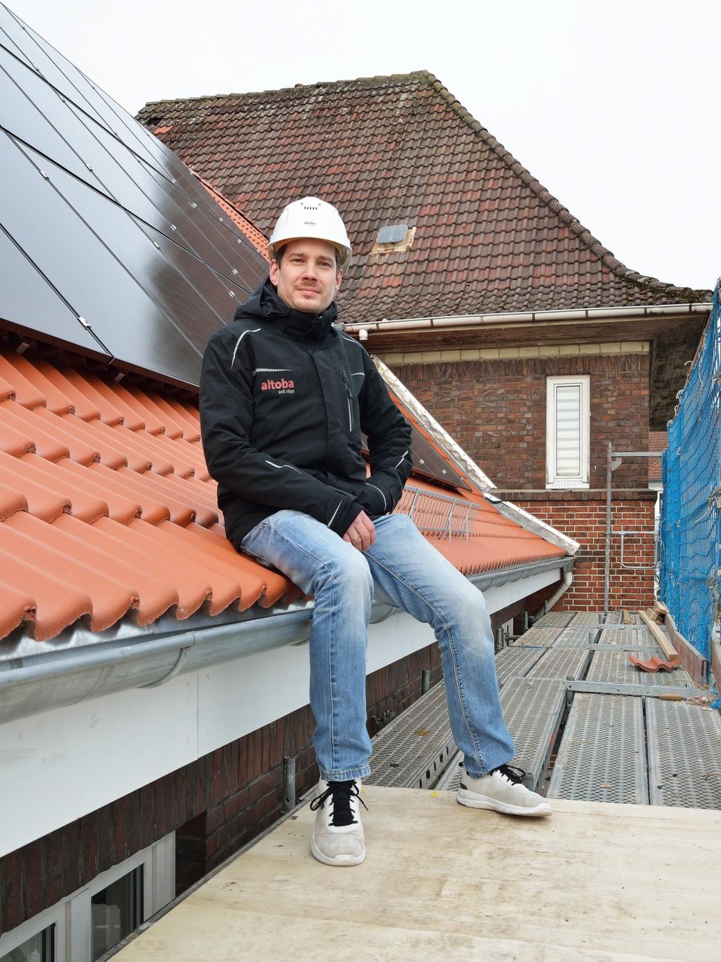 Ein Mann mit weißem Bauhelm und altoba-Jacke sitzt auf einem Gerüst am Rand eines Ziegeldachs mit installierten Solarpaneelen; im Hintergrund sind weitere Dächer und ein Sicherheitsnetz zu sehen.
