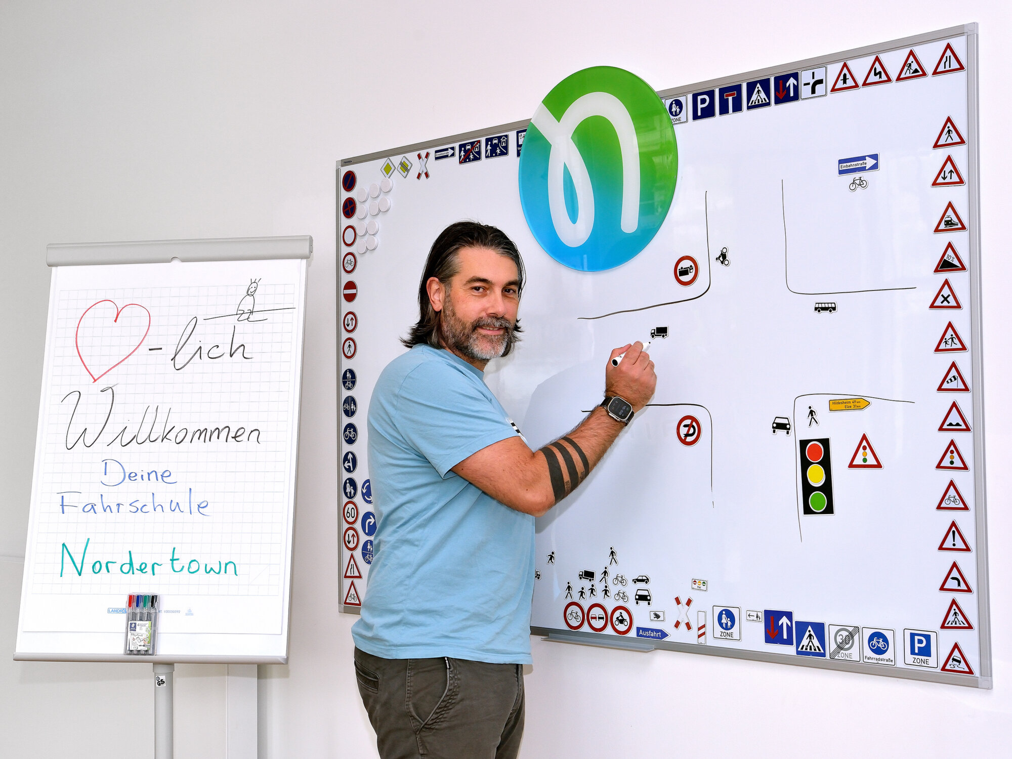 Ein Mann steht vor einem Whiteboard mit Verkehrsschildern und Straßensymbolen und zeichnet eine Kreuzungsszene; daneben steht ein Flipchart mit der handschriftlichen Begrüßung „Herzlich Willkommen – Deine Fahrschule Nordertown“.