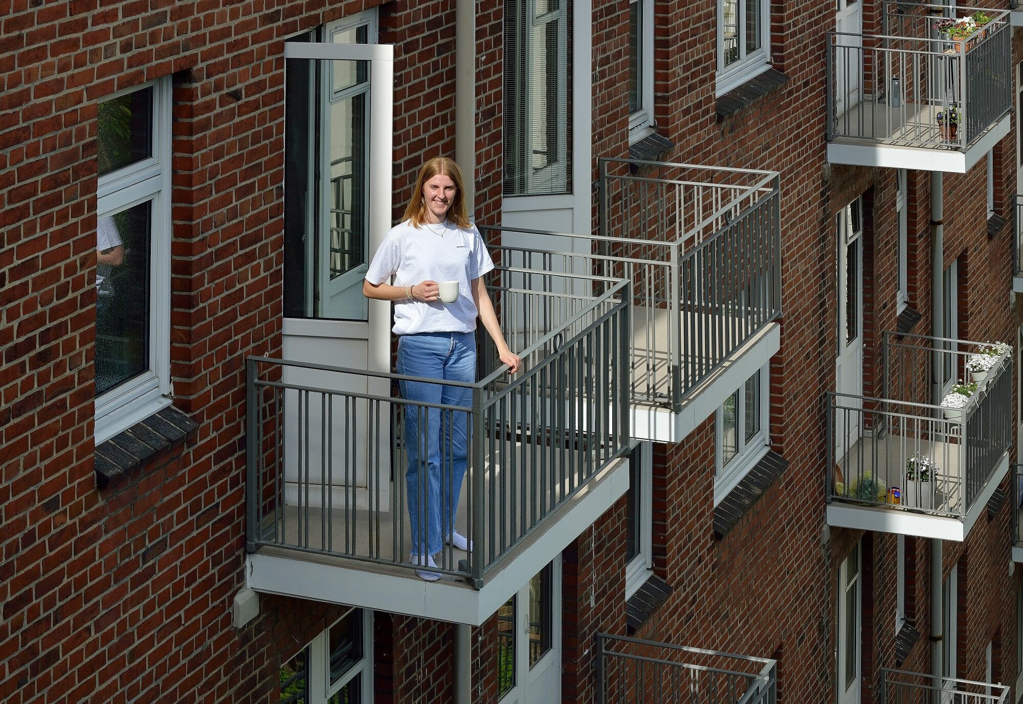 Eine junge Frau steht mit einer Tasse in der Hand auf einem kleinen Balkon eines mehrstöckigen Wohngebäudes aus rotem Klinker; mehrere ähnliche Balkone mit Blumenkästen sind an der Fassade zu sehen.