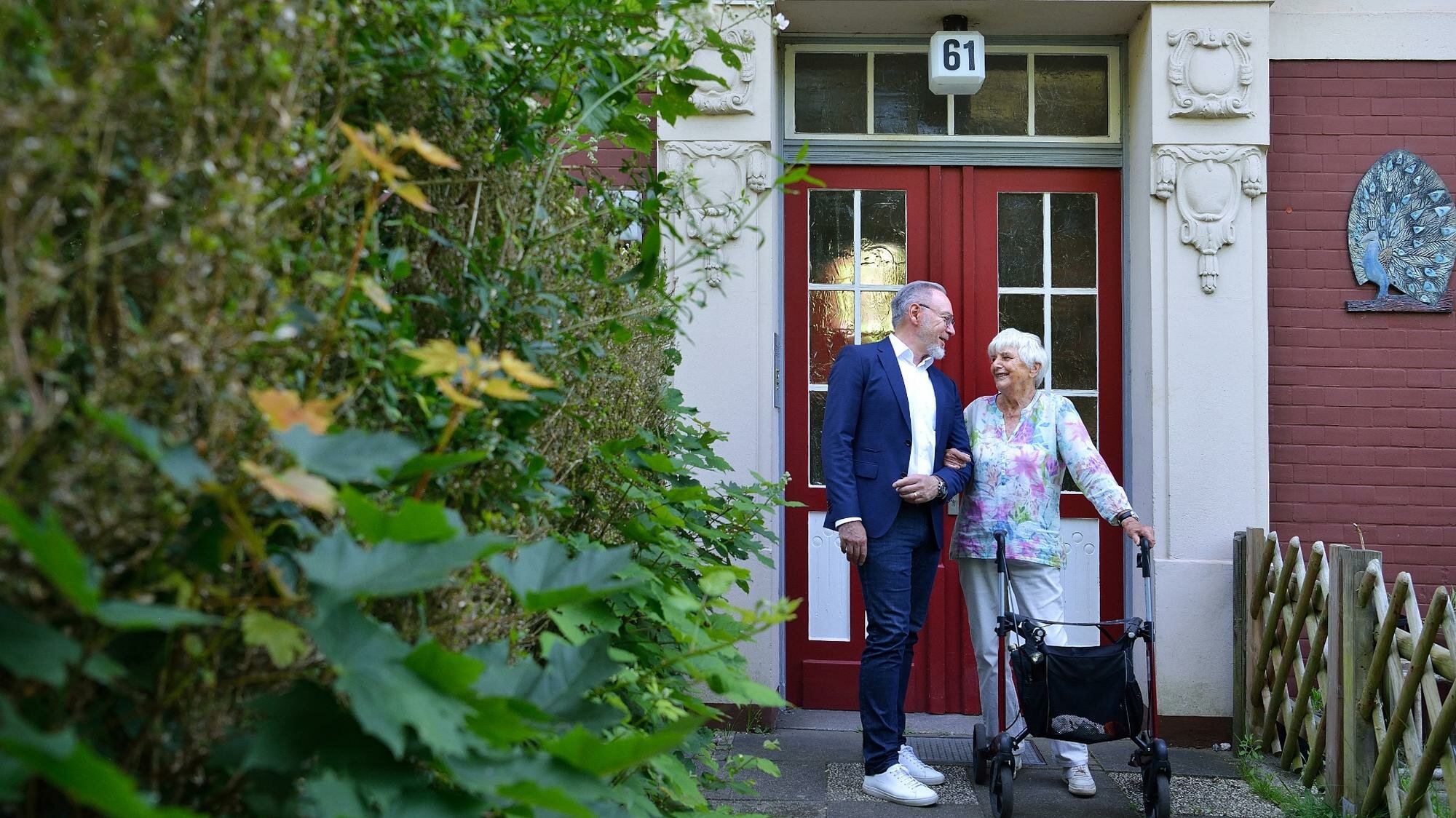 Ein Mann im Anzug und eine ältere Frau mit Rollator stehen gemeinsam lächelnd vor einem Hauseingang mit der Hausnummer 61 und unterhalten sich.