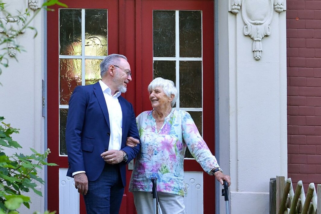 Ein Mann im Anzug und eine ältere Frau mit Rollator stehen gemeinsam lächelnd vor einem Hauseingang mit der Hausnummer 61 und unterhalten sich.