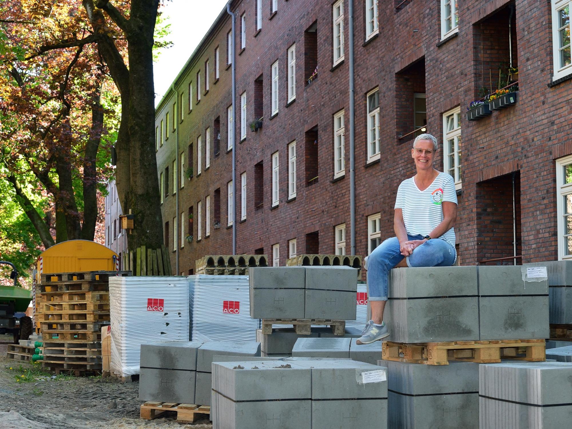 Eine Frau mit kurzen Haaren, Brille, weiß gestreiftem T-Shirt und Jeans sitzt lächelnd auf gestapelten Pflastersteinen vor einer roten Klinker-Wohnhausfassade; im Hintergrund stehen Bäume und Baumaterialien auf einer Baustelle.