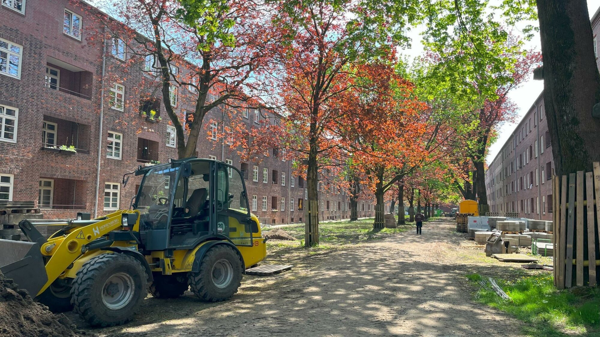 Blick auf eine Baustelle zwischen zwei mehrgeschossigen Backsteingebäuden mit typischer Altbauarchitektur; im Vordergrund steht ein gelber Radlader, entlang des unbefestigten Weges sind Bäume mit rot und grün belaubten Kronen sowie Baumaterialien und Absperrungen zu sehen.
