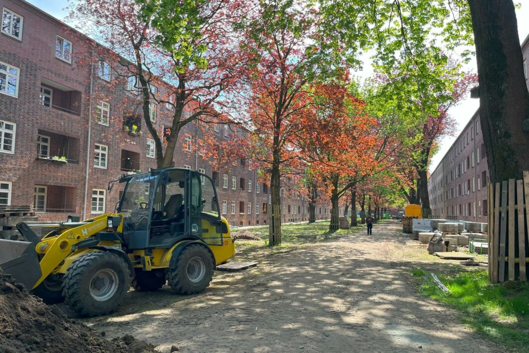 Blick auf eine Baustelle zwischen zwei mehrgeschossigen Backsteingebäuden mit typischer Altbauarchitektur; im Vordergrund steht ein gelber Radlader, entlang des unbefestigten Weges sind Bäume mit rot und grün belaubten Kronen sowie Baumaterialien und Absperrungen zu sehen.