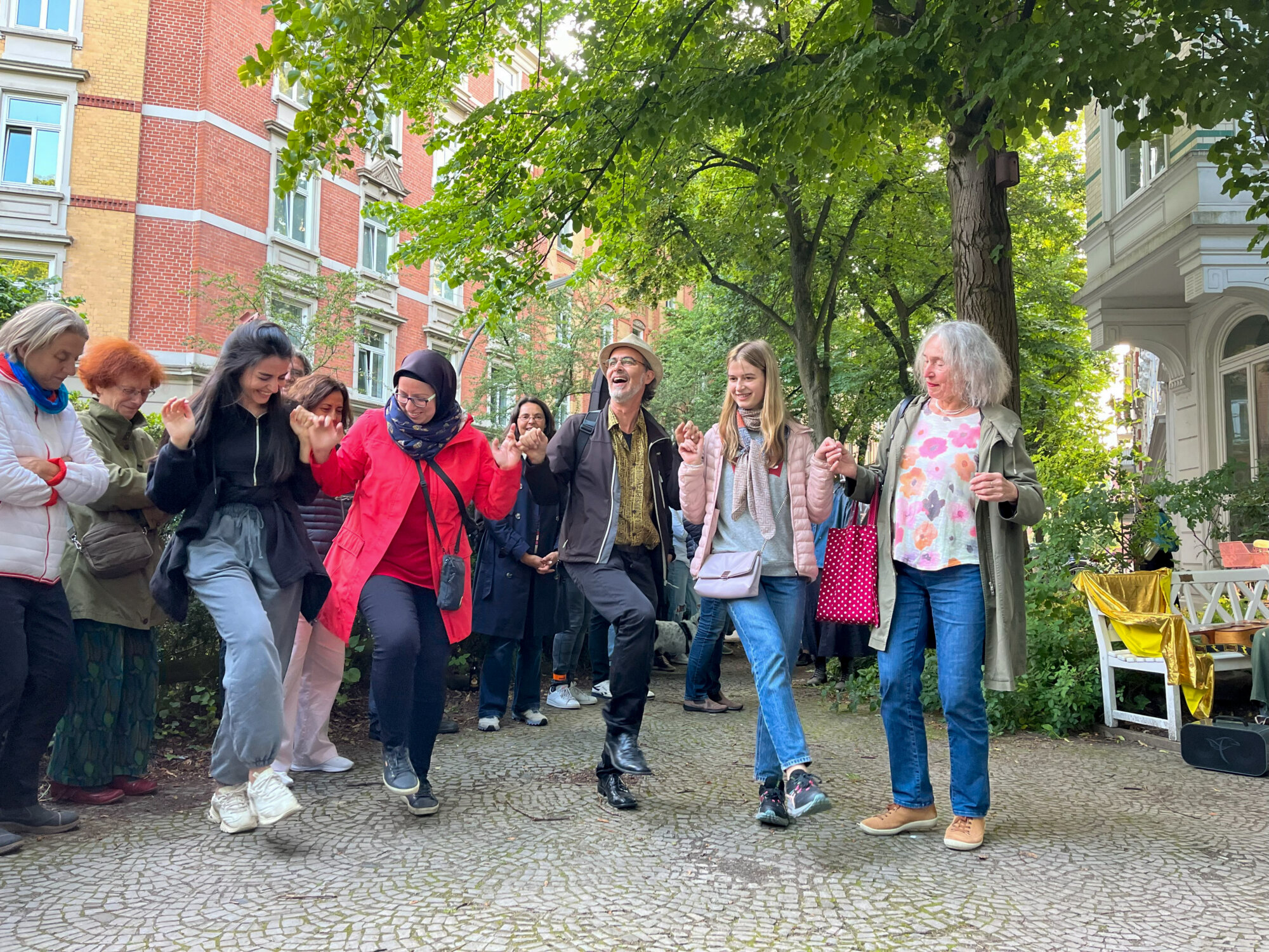 Mehrere Erwachsene tanzen fröhlich in einer Reihe auf einem gepflasterten Weg unter Bäumen in einem Wohnquartier mit Altbaufassaden; sie halten sich an den Händen und werden von einer Gruppe beobachtet.