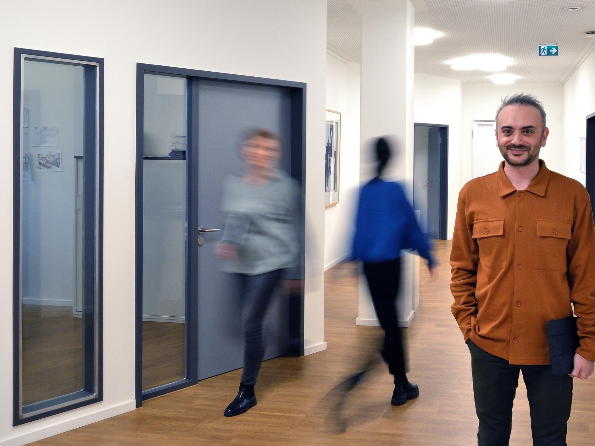 Ein Mann mit Tablet steht in einem hellen Büroflur mit Holzfußboden; im Hintergrund gehen zwei unscharf dargestellte Personen an geschlossenen Bürotüren vorbei.