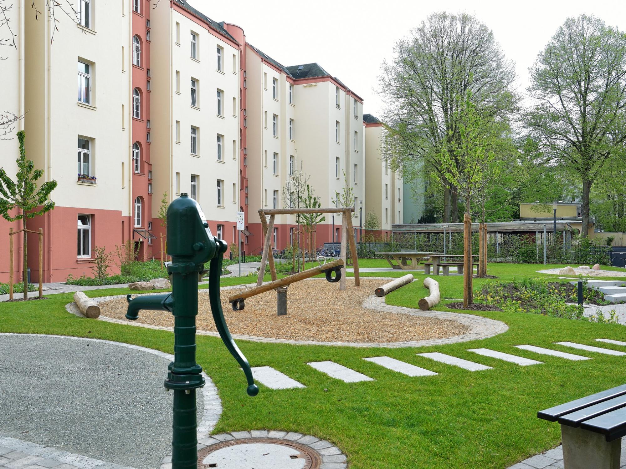 Innenhof einer Wohnanlage mit grünem Spiel- und Aufenthaltsbereich; im Vordergrund eine historische Handpumpe, dahinter ein Spielplatz mit Wippe und Schaukel aus Holz sowie Sitzbänken und Picknicktisch; umgeben von mehrstöckigen, hellen Wohngebäuden mit roten Sockeln.